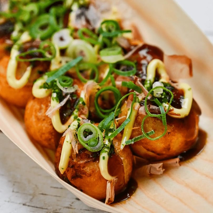 Close-up of golden-brown takoyaki topped with green onions, mayonnaise, and sauce in a wooden boat.